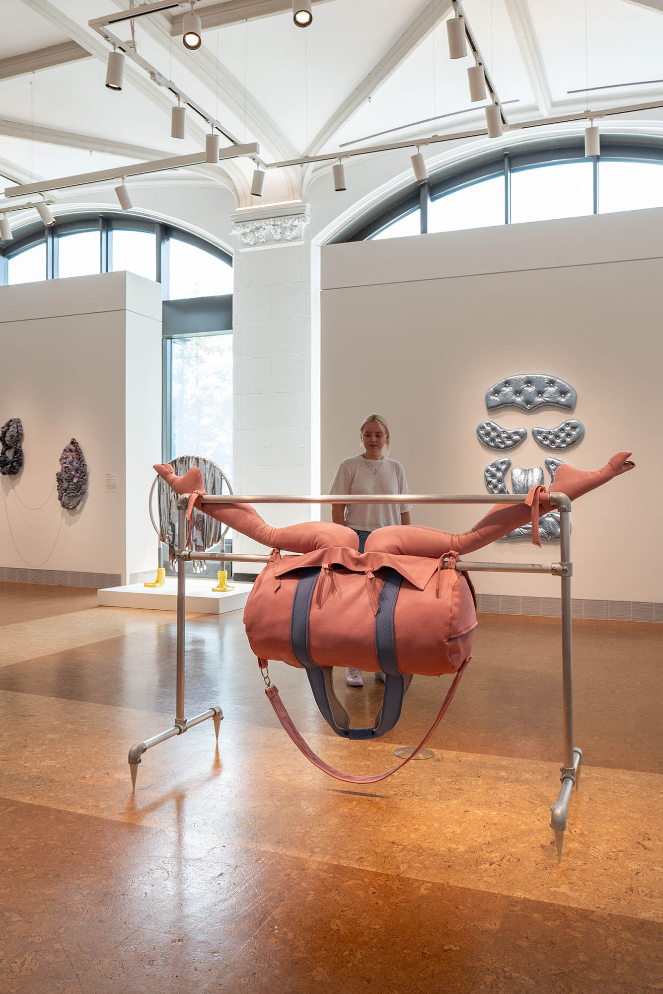 Photo: A salmon-colored stuffed duffle bag, hanging on metal rods so that it is suspended in the air. Two, large, stuffed leg-type appendages stretch out from left to right on top of the bag. The artwork is amongst the middle of a gallery floor, surround by smiliar art pieces. There is a short woman standing behind the piece, observing.