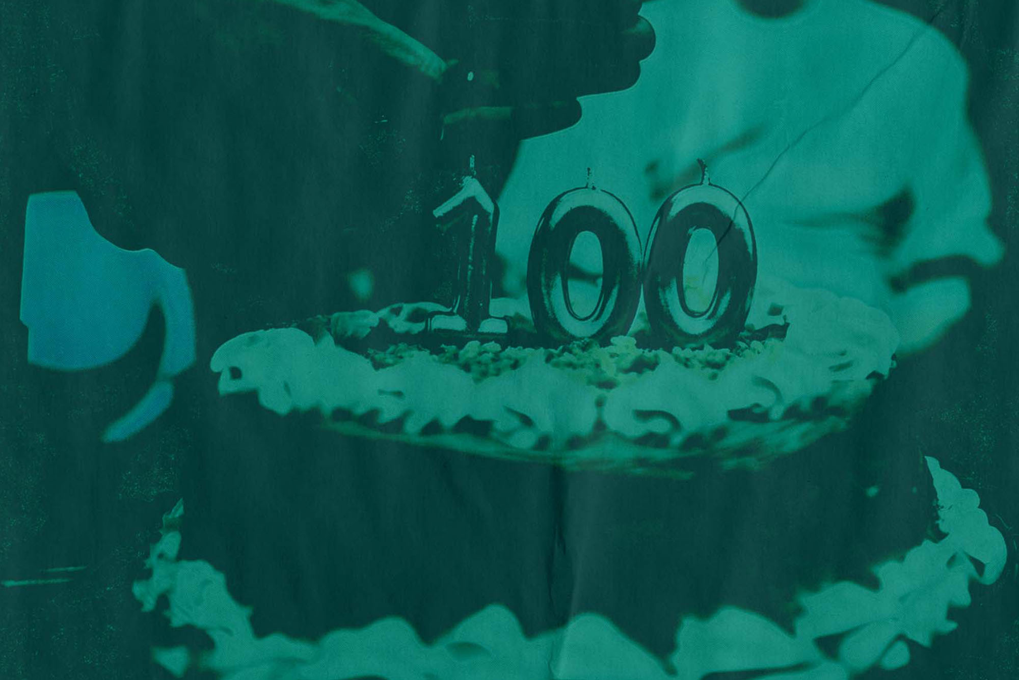 Image of a birthday cake with silver candles that read "100" on top. A hand with a lighter goes to light the birthday candles. A person is seen seated, blurred, in the background.