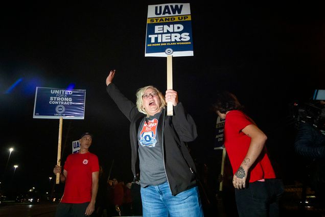 Photo: A white woman wearing a black jacket, tee, and jeans, shouts and raises a sign that reads "UAW Stand Up, End Tiers, No more second class workers".