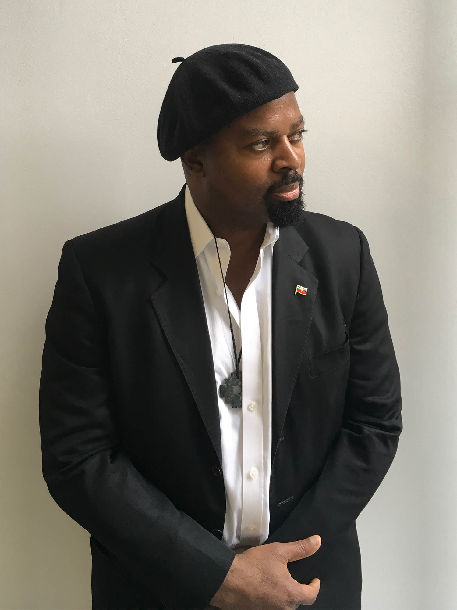 Photo: A black man in a beret and a collared shit with a suit jacket poses for a formal photo