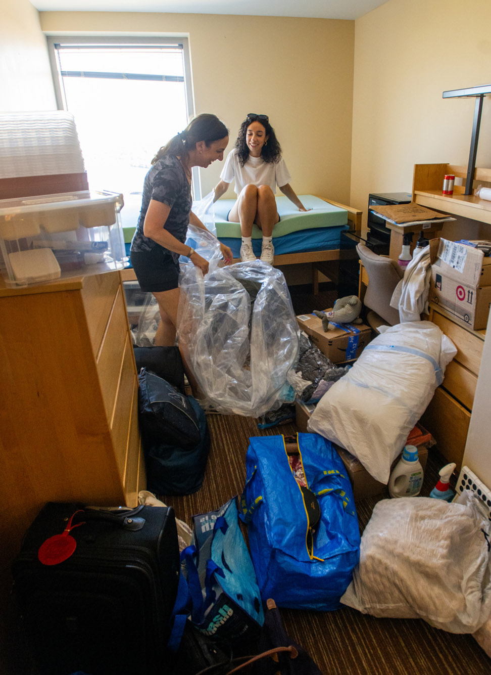 Photo: Two woman laugh at all the stuff pulled amongst the dorm room. One is on the bed, the other is in the middle of folding a plastic bag covering back up. Across the floor are various amounts of duffle bags, bags, open boxes, bins, plastic boxes, and more.