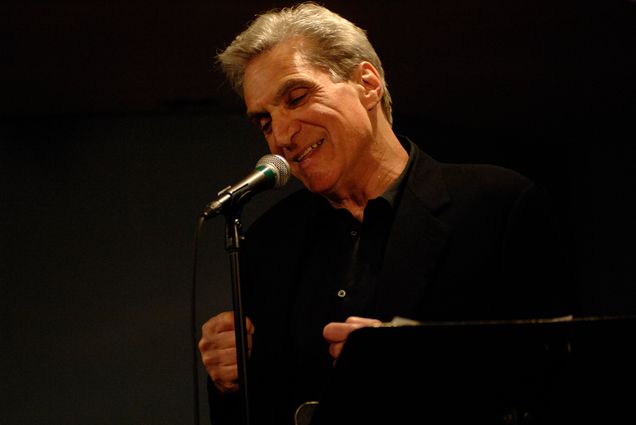 Photo: Poet Robert Pinsky, a white man with a full head of gray hair, stands at a mic with a black button down as he reads his poetry. A single spotlight illuminates him.