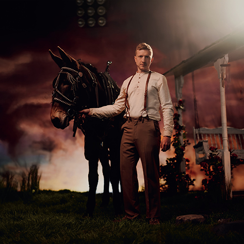 Album cover of Tyler Childers' "Rustin’ in the Rain". cover shows Childers, a white man wearing suspenders, brown pants, and long-sleeved white shirt, standing in a field next to a dark brown horse. The scene is dark and moody.
