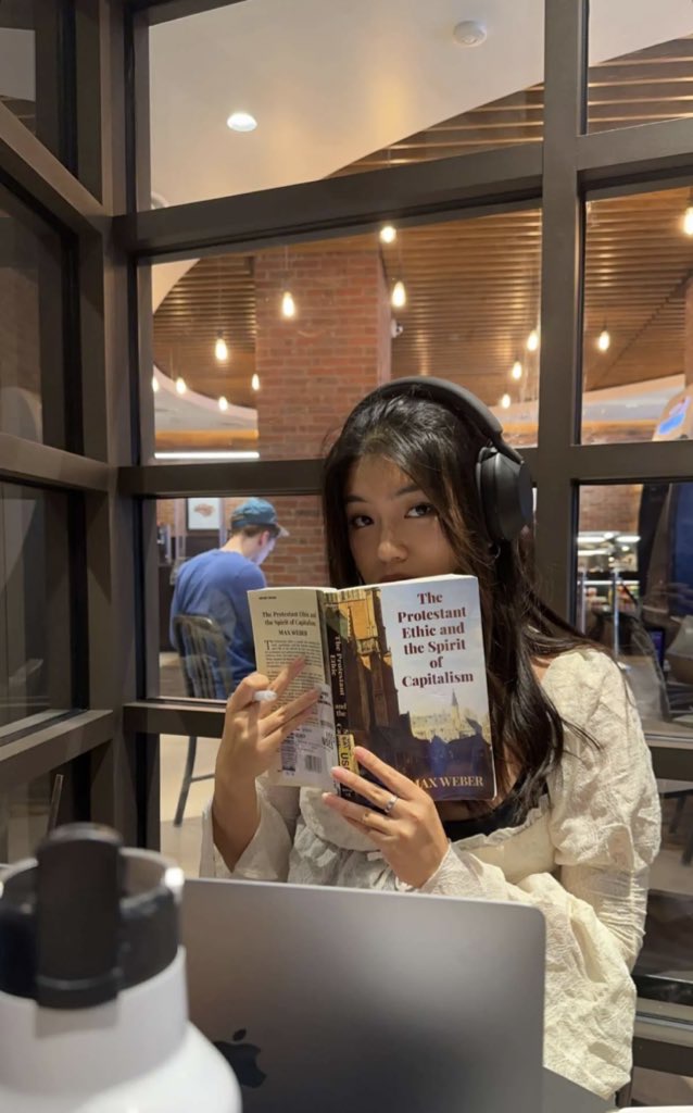 This is a photo of Robin Kim (COM'24) reading  a book in a coffee shop. She has brown hair and headphones on and the book is covering half of her face. In front of her, there is an open laptop and a water bottle. The book she is reading is titled  The Protestant Ethic and the Spirit of Capitalism. She also has a pencil in one hand. She is looking right at the camera and is in front of a large set of windows. 