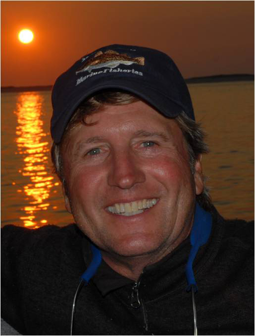 Photo: Greg Skomal, a white man wearing a black cap smiles widely as he poses in front of a large body of water at sunset.