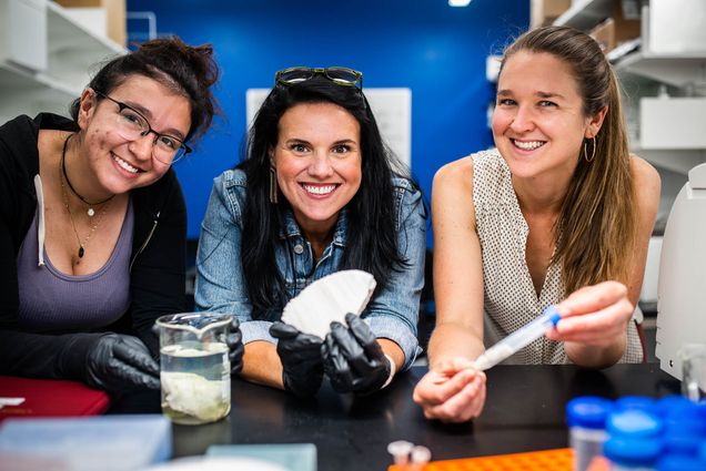Photo: Group photo of Assistant Professor of Biology, Sarah Davies (center) and PhD student, Maria Valadex Ingersoll (CAS 23) and Dr Hannah Aichelman (right) (CAS 23). The three women pose in a lab as Davies holds up a sample of bleached coral and another sample of algae rests on the lab tables in front of Ingersoll.