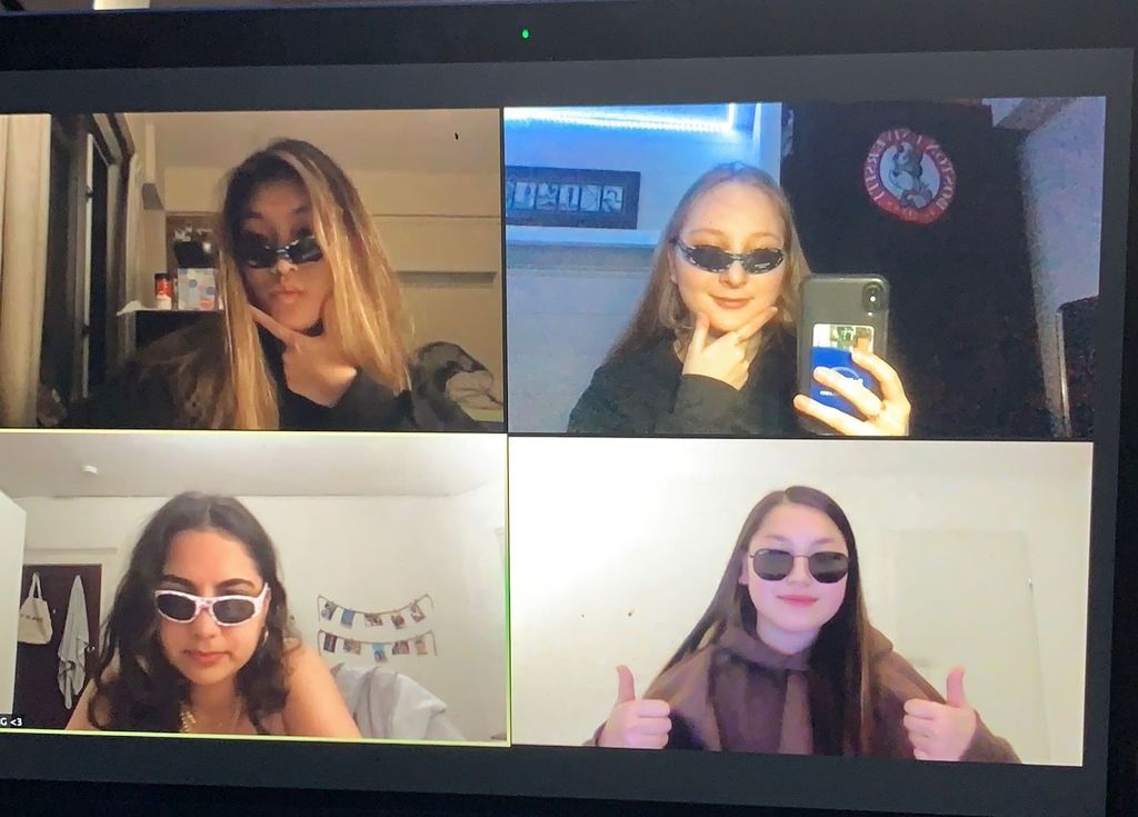 This is a screenshot from a group Zoom call. There are four individuals on the call, all wearing black sunglasses. They are all girls and they are in casual attire. They have straight faces on and the individual in the upper right corner has her phone on the screen, as if she was taking a picture. The individual to the left of her has her hand below her chin. The individual directly below her has a straight face on, looking straight at the camera. The person to the right of her has two thumbs up with a straight face on as well. 