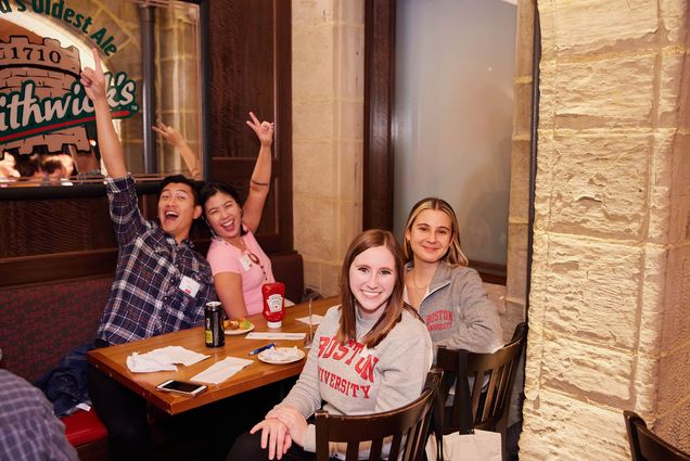 Photo: Four individuals sit at a table. The two on the farthest side threw up peace signs and big smiles. The two on the nearest side, the side in the foreground of the photo, sit in their chairs, turned around and smile at the camera.
