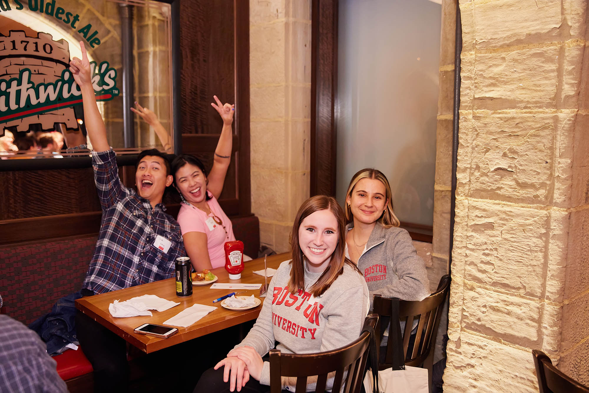 Photo: Four individuals sit at a table. The two on the farthest side threw up peace signs and big smiles. The two on the nearest side, the side in the foreground of the photo, sit in their chairs, turned around and smile at the camera.