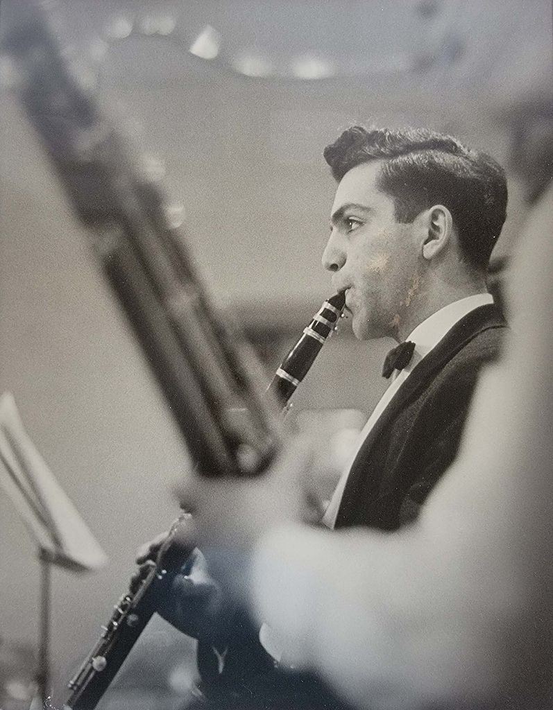 Black and white photo taken of Ed as a young man, performing the clarinet at BU. He wears a tuxedo and his dark hair is slicked back. He looks at music on a music stand and another wind player is seen in the foreground.