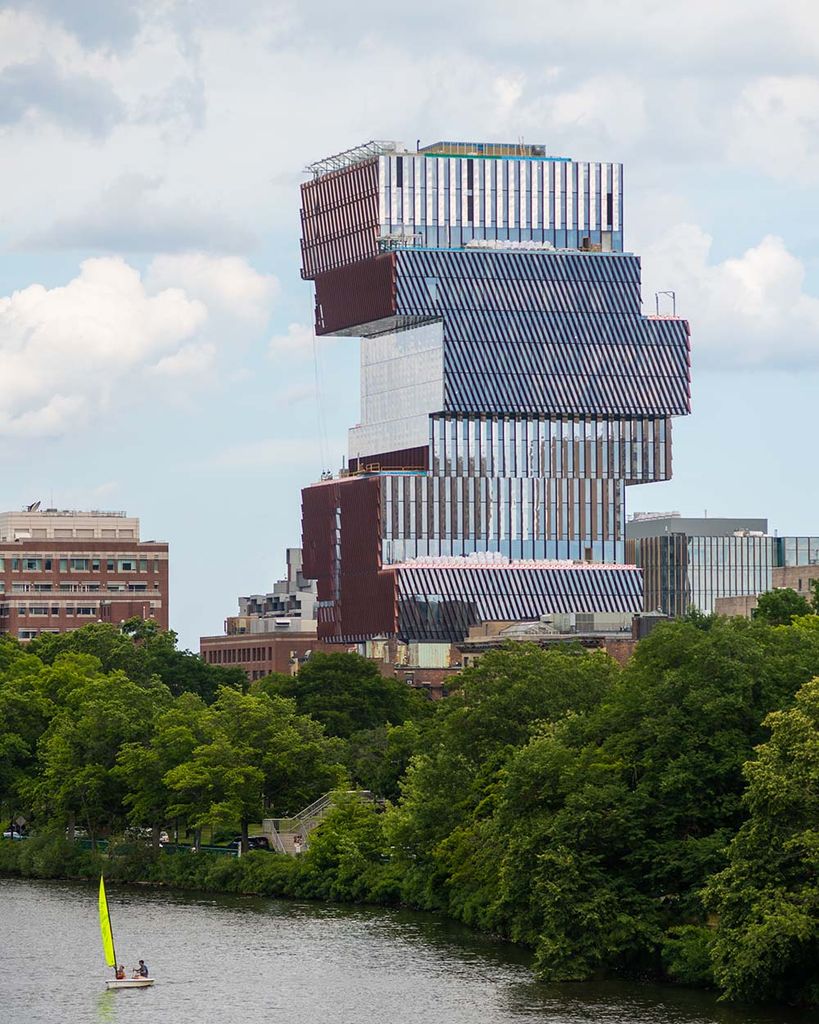 Photo of the Boston skyline as seen from the BU bridge. The reflective blue and reddish Center for Data Science features prominently at right. Bright green summer trees line the Charles River and the sky is a bit overcast. On the river, a small sunfish sailboat with a yellow-green sail is seen.