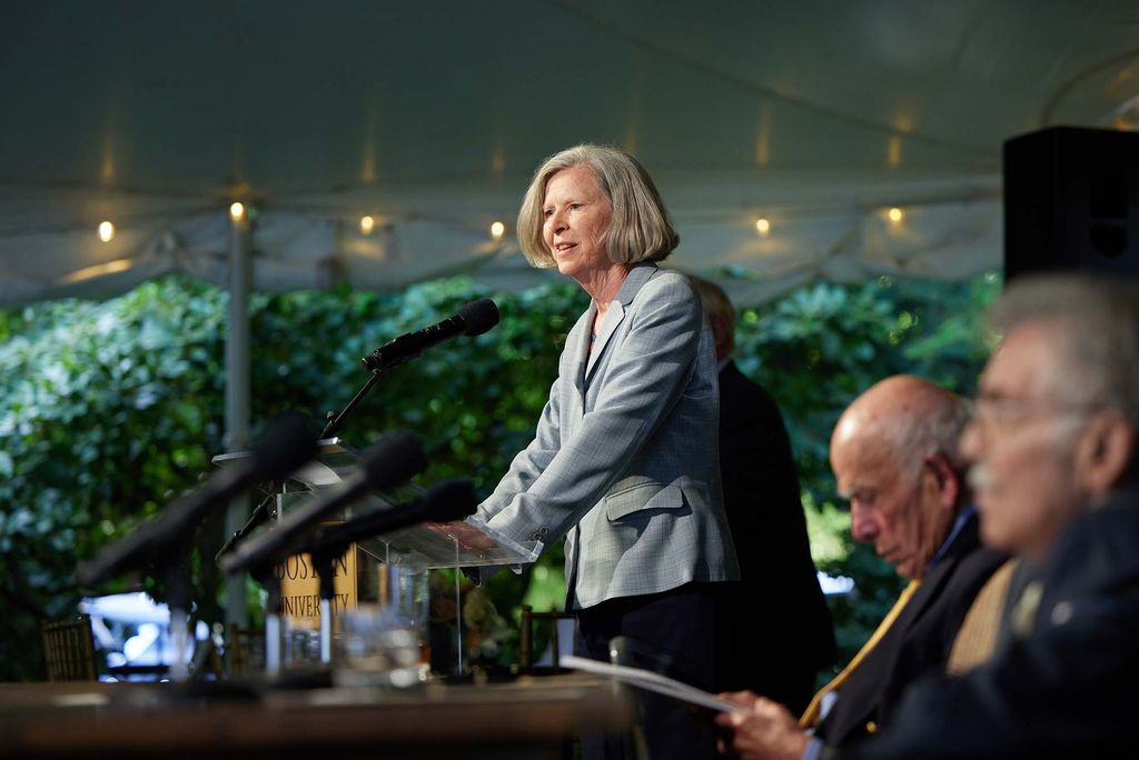 Photo of Karen Antman, dean of the School of Medicine and provost of the Medical Campus, speaking at a wooden podium. Avram and Edward are seen blurred, at right, in the foreground. Antman is a White woman with ear-length blonde hair who wears a pale blue pant suit.