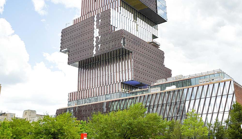 Photo of students taking their lunch breaks on the lawn in front of College of Communication building with the newly built Center for Data Sciences towering in the background. The building resembles a stack of books, and is very reflective. Students sit in white and green Adirondack chairs; the lawn and trees along the sidewalk are bright green. The sky is light blue and filled with large white clouds.