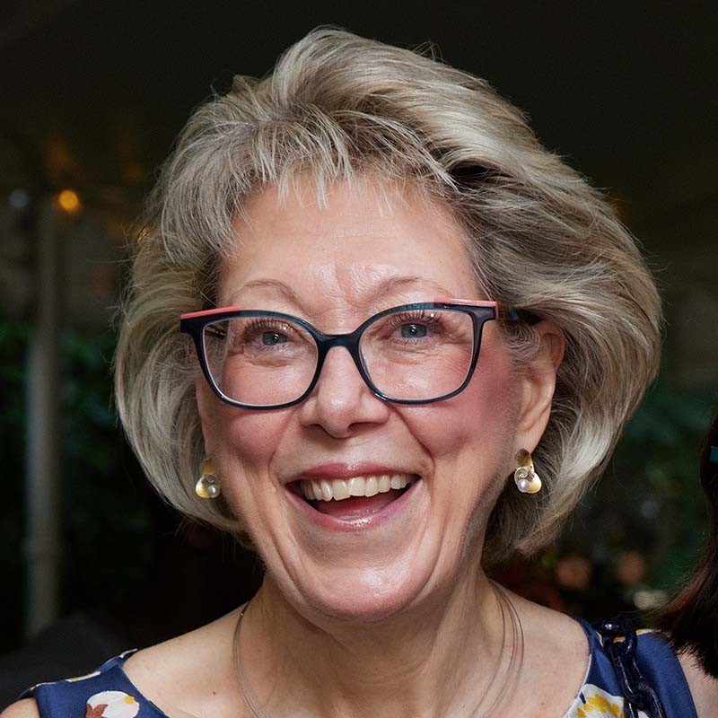 Pamela Avedisian, a White woman with blonde hair and glasses who smiles. She wears a dark blue floral dress and gold earrings.