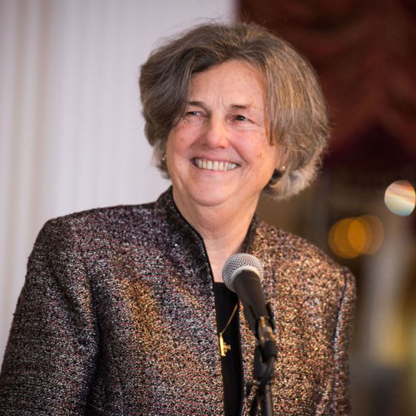 A photo of Phyllis Zagano in a brown blazer jacket with a microphone on a podium in front