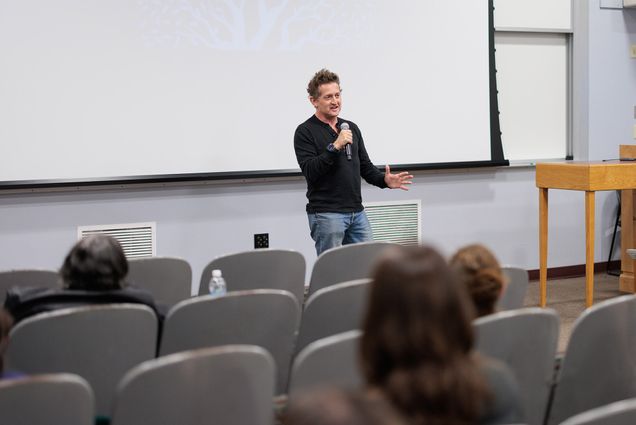 Photo: A man holds a microphone while speaking to an audience in a large auditorium