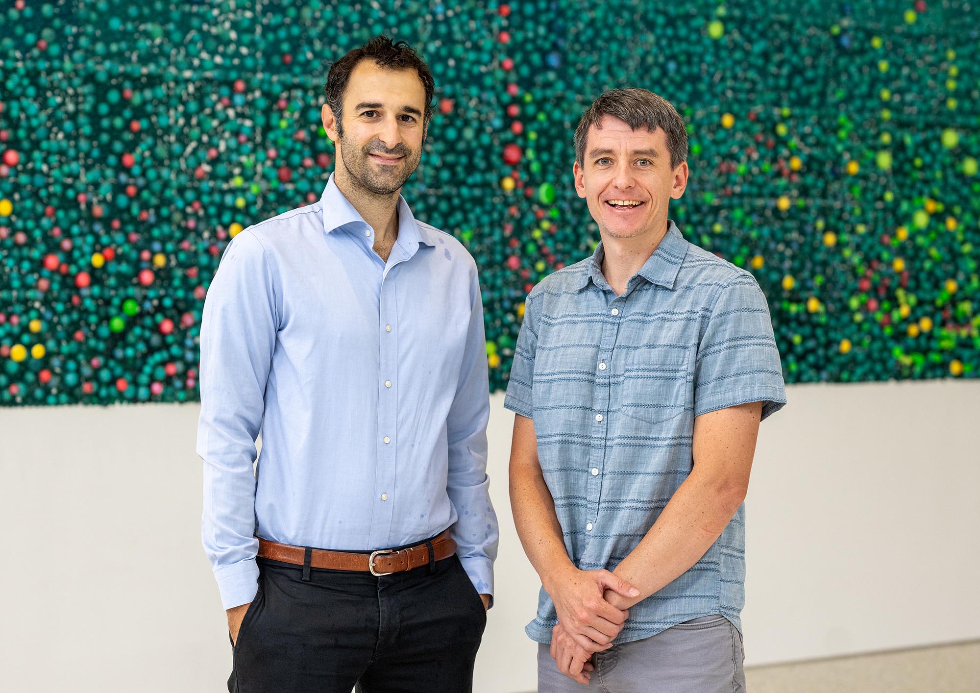 Photo: Brian Cleary, right, and Michael Economo stand and pose for a photo together. Cleary is a white man wearing a plaid light blue collared shirt and grey slack. Economo is a white man wearing a light blue, log sleeved collared shirt and black slacks. They pose in front of a green foliage wall.
