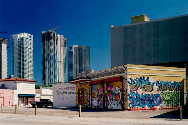 Photo: an urban downtown scene of the Miami cityscape is shown. A small building covered in graffiti is juxtaposed in the foreground against new, tall glass buildings behind it.