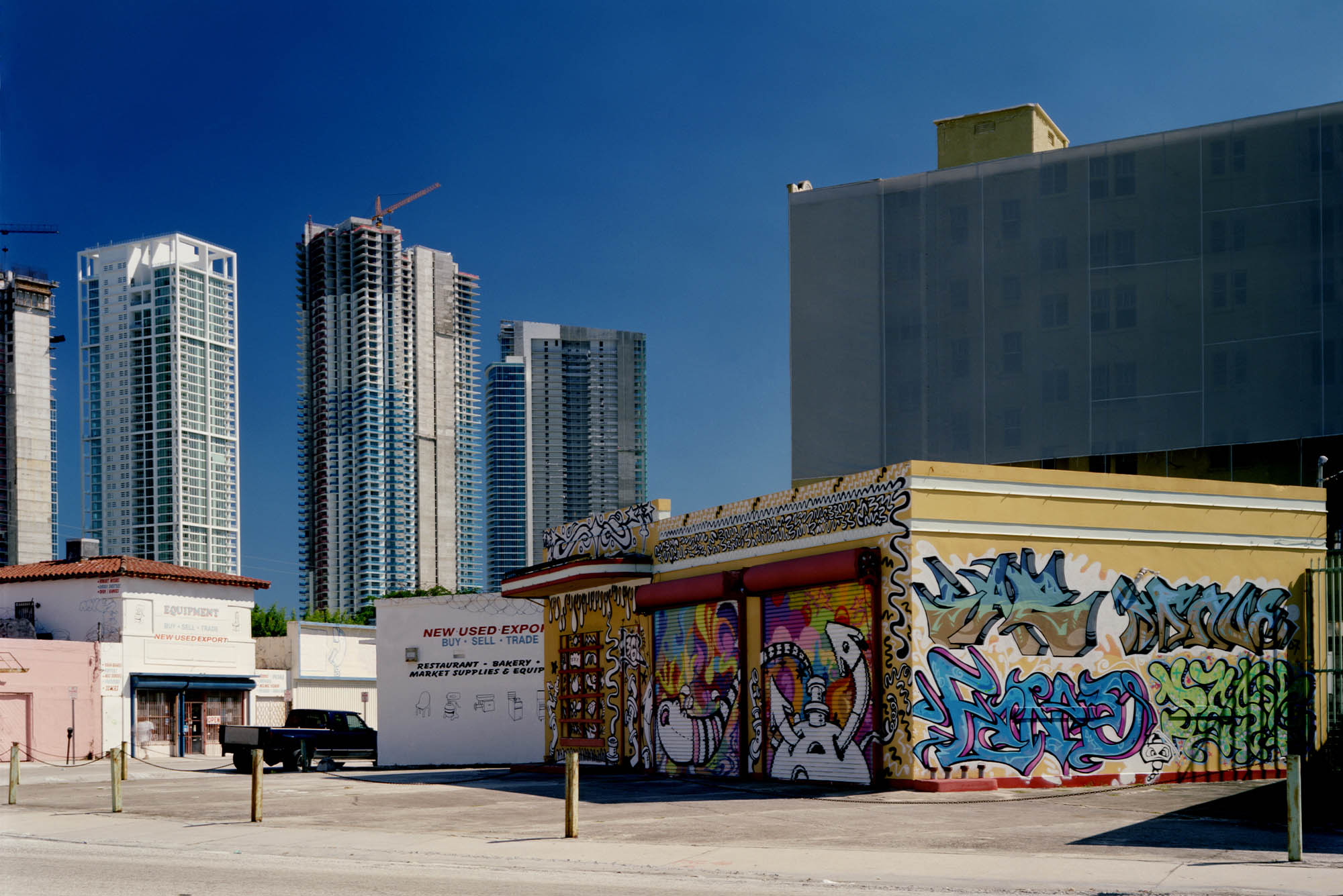 Photo: an urban downtown scene of the Miami cityscape is shown. A small building covered in graffiti is juxtaposed in the foreground against new, tall glass buildings behind it.