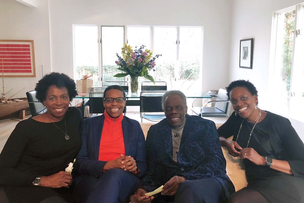 Photo: A family of four Black people smile and pose for a photo together. They sit on a cream couch and sit in a brightly lit living room.
