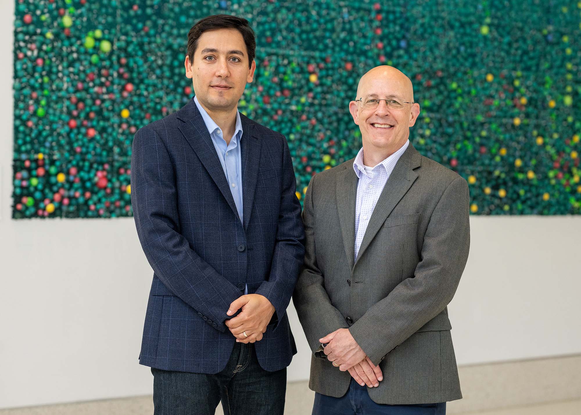 Photo: Hadi Nia, left, and Jospeh Mizgerd stand and pose for a photo together. Nia is a tan man wearing navy blue blazer and slacks. Mizgerd is a white man wearing a light blue collared shirt, grey blazer, and grey slacks. They pose in front of a green foliage wall.