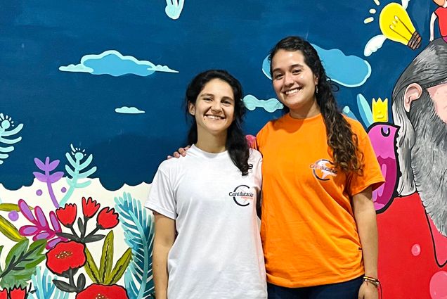 Photo: Two young adults stand side by side in front of a large colorful mural