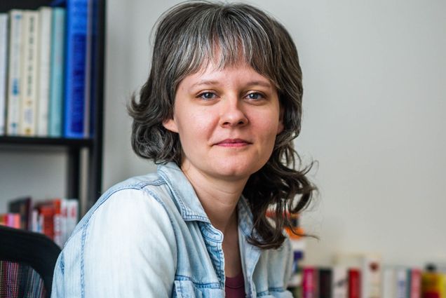 Photo: A white woman with dark grey hair and lightly silver bangs smiles and poses in her studio. She sits in a black office chair and wears a burgundy shirt and unbuttoned blue collared shirt.