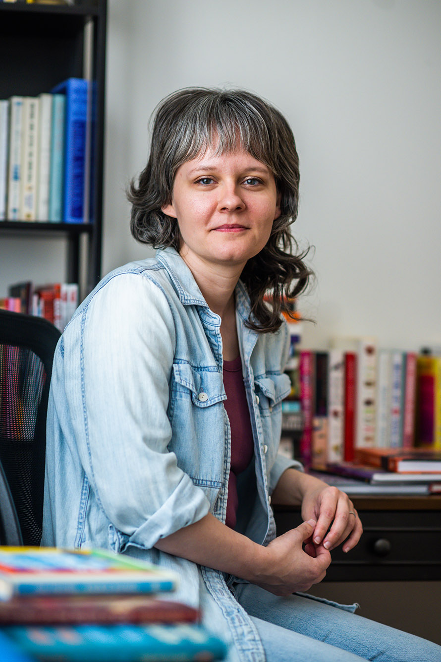 Photo: A white woman with dark grey hair and lightly silver bangs smiles and poses in her studio. She sits in a black office chair and wears a burgundy shirt and unbuttoned blue collared shirt.