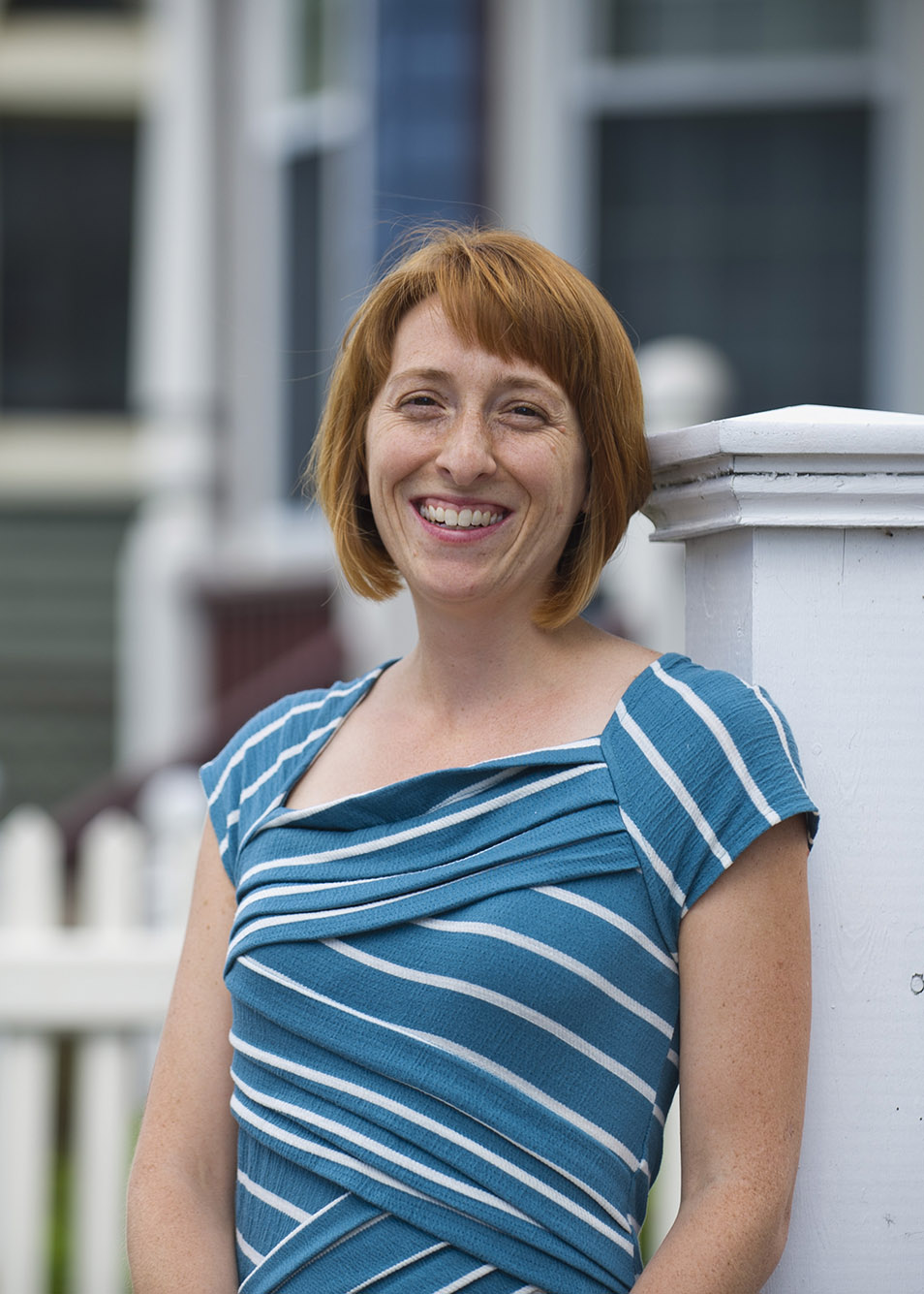 Photo: Japonica Brown-Saracino, a white woman with auburn hair and wearing a blue and white blouse, smiles as she poses next to a white fence in a suburban neighborhood.