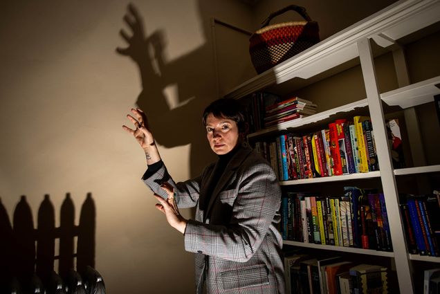Photo: Maia Gil'Adi, a white woman with short, dark brown hair and wearing a grey suit, makes a zombie pose in front of light in a dim room. Her pose creates a creepy shadow behind her, emphasized by the lighting.