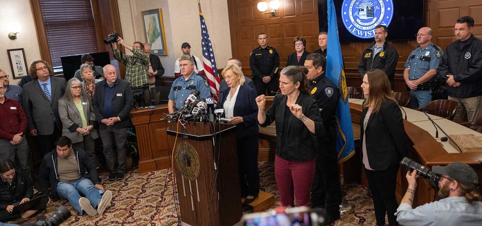 Photo: wide shot of Maine Governor Janet Mills speaking during a press conference about the mass shooting on October 26, 2023 in Lewiston, Maine. A white woman in business attire stands at a wooden podium front and center of the room. Behind her, police officers and officials stand. To her right, an ASL interpreter signs. In front of her, various journalists and photographers write, take photos, and listen.