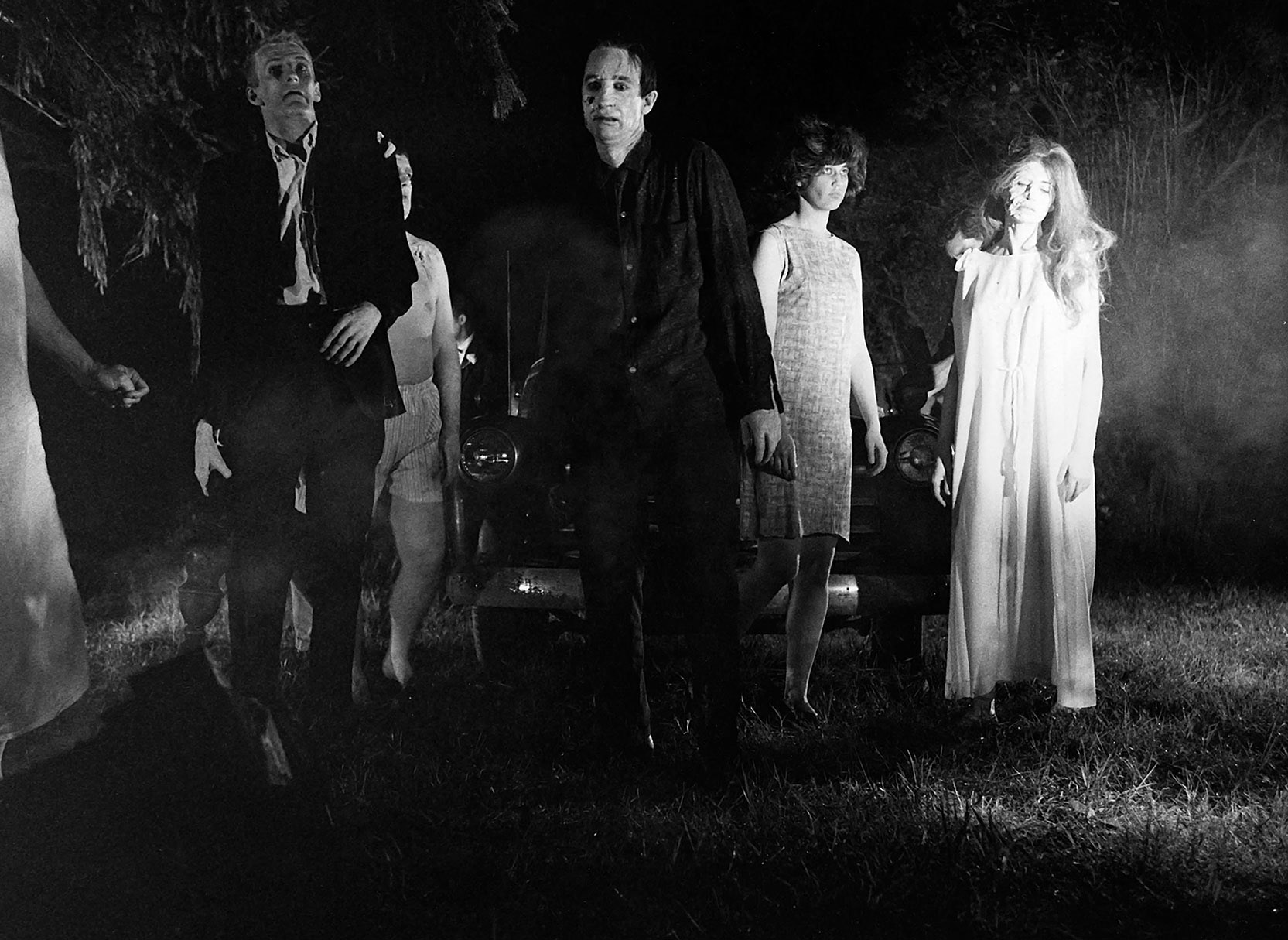 Image: Black and white screenshot of a scene from the movie "Night of the Living Dead". Various zombies are shown listlessly moving in a cemetery.