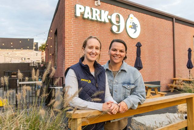 Photo: Emily Gusse, a young white woman wearing a grey long-sleeved shirt and black puffer vest smiles and poses next to Tess Kohanski, a white woman wearing a grey sweater. They both pose in front of a large brick building with a large sign that reads "Park-9".