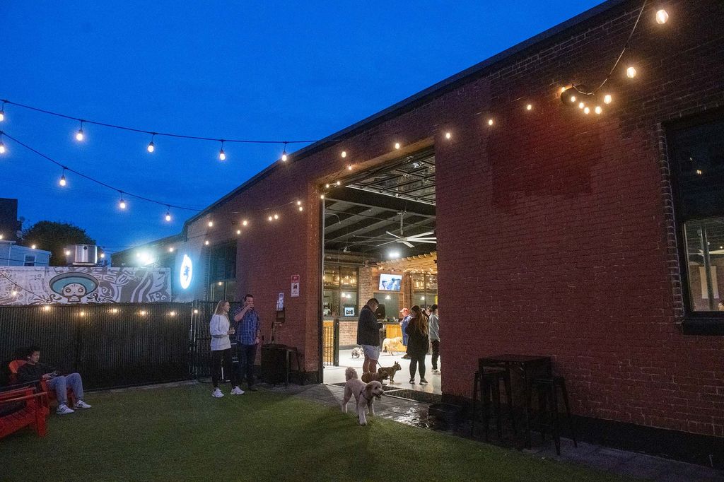 Photo: Exterior of Park-9 dog bar in the evening. A brick building is shown with large open doors. Dogs and people can be seen inside at string lights light up the dusky night.