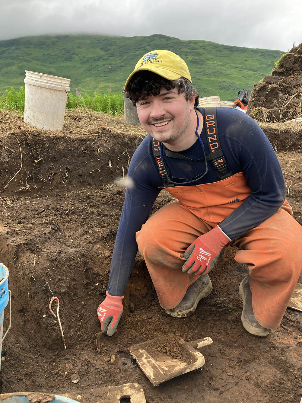 Photo: Trevor Lamb, a young white man wearing a navy blue long-sleeved shirt, yellow baseball cap, and orange overalls and gloves, smiles as he bends down in a small archeology dig site.