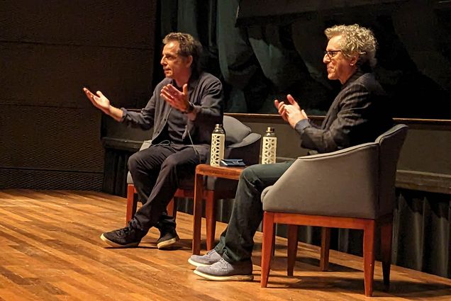 Photo: Photo: Ben Stiller, a white man with graying hair wears a dark, dusty blue button up with similar color shirt and slacks. He sits in a plush chair on a wooden stage, hands up, talking. Next to him, in another plush chair, is Jeff Kahn, another white man with a blue button up, a pinstripe blazer, and the same color pants as Stiller. He is smiling as he talks, faced in the same direction towards the audience as Stiller is.