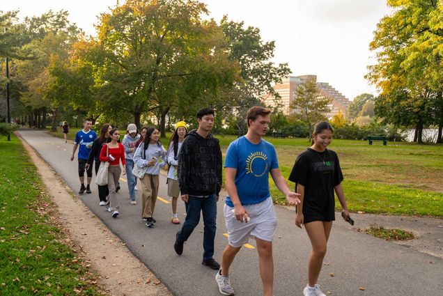 Photo: A group of young college students walk on a paved sidewalk in groups of two. The greenery around them is bright, with hints of fall in the leaves.