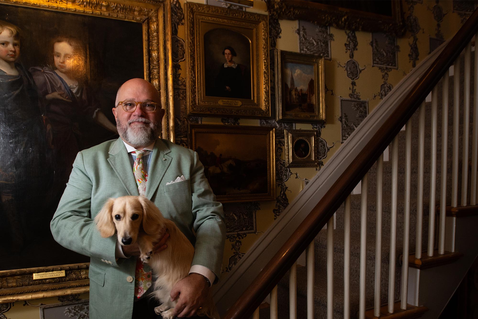 Photo: A bald man in a suit holds a dachshund while standing on an ornate wooden staircase in front of many old time portrait paintings