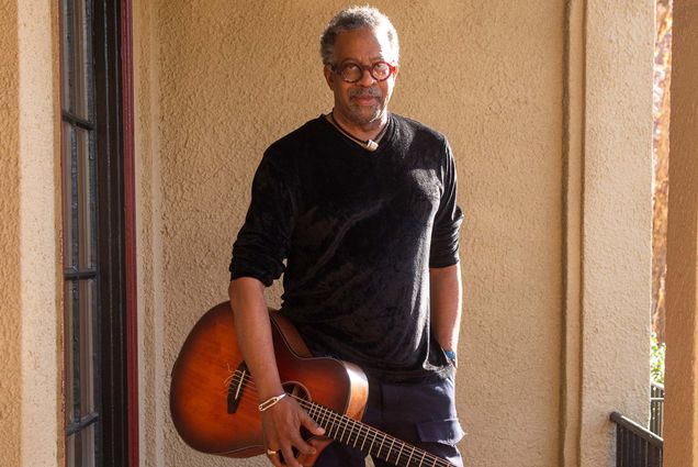 Photo: Bill Banfield, A Black man short graying hair wearing red glasses, a black long sleeved shirt with sleeves rolled up to his elbows, and black pants, poses as he holds his guitar under one arm. He stands at the exterior of a Spanish style building deck as the sun shines on him.