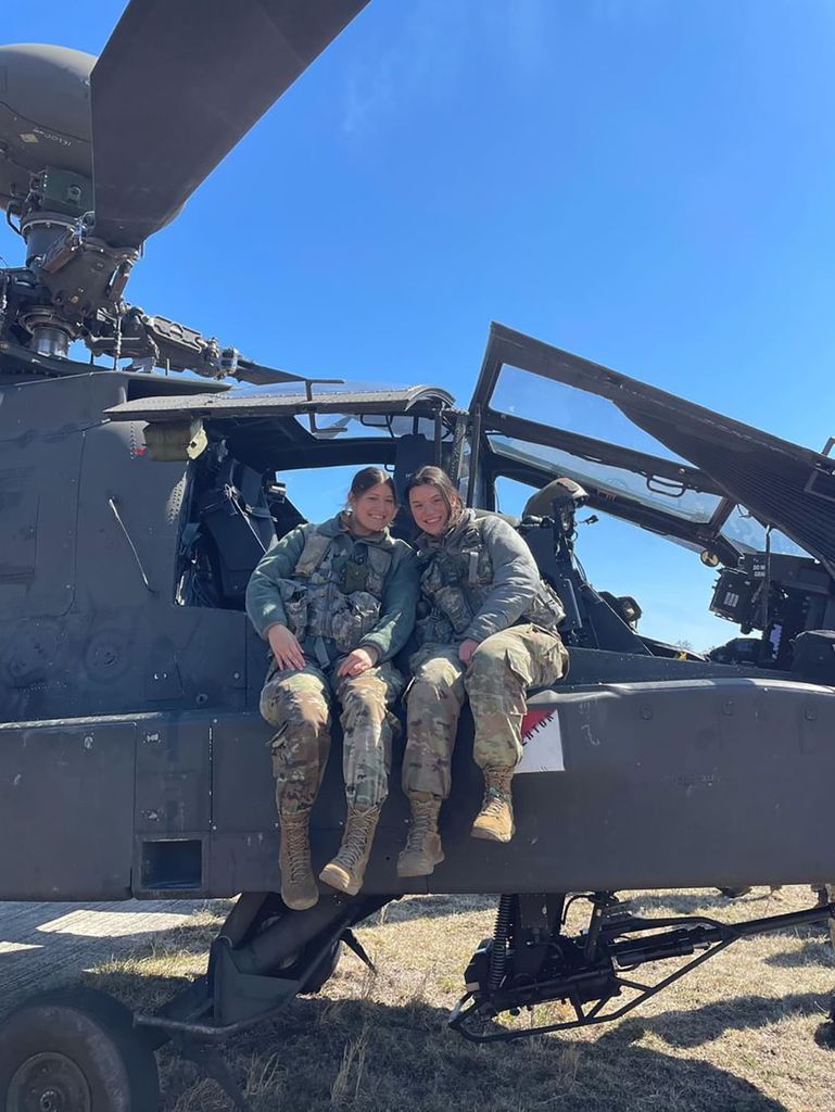 Photo: Two young women wearing Army ROTC fatigue gear sit outside the cockpit of a military helicopter.