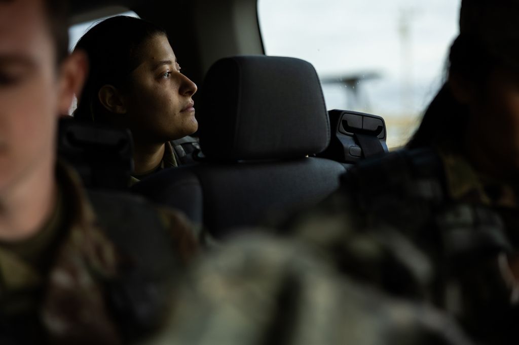 Photo: Dharma Cooper, a young woman with hair slicked back into a tight bun, looks to to the right towards the window of the van she sits in. She wears US Army ROTC gear, as does the blurred people in the foreground.