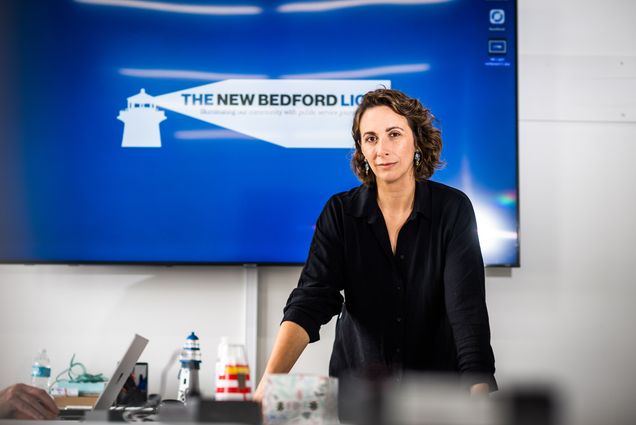 Photo: Daniela Melo, a white woman with short, curly brown hair and wearing a black, long-sleeved collared shirt leans forward against a white counter with various lighthouse and editorial paraphernalia on it. Behind her a large tv screen displays the blue lighthouse logo of "The New Bedford Light"