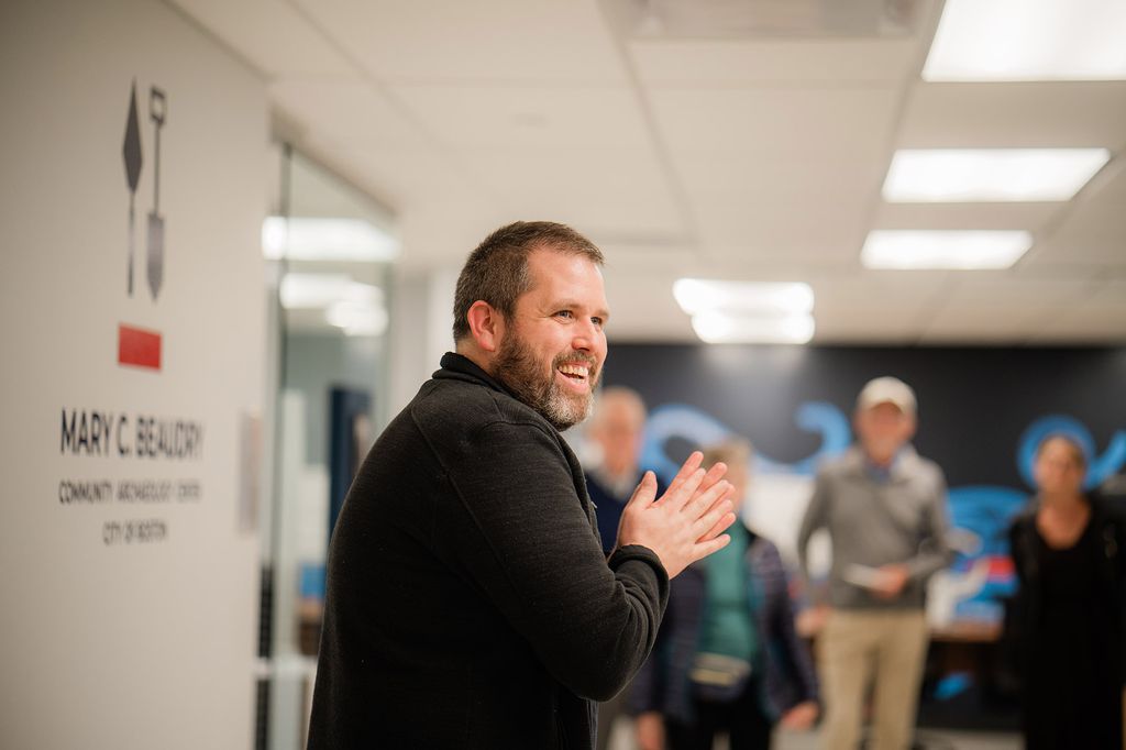 Photo: Joe Bagley, a white man wearing a black long-sleeved top, smiles and holds his hands together as he addresses an unseen audience. He stands in front of a wall that reads "Mary C. Beaudry Community Archaeology Center, City of Boston".