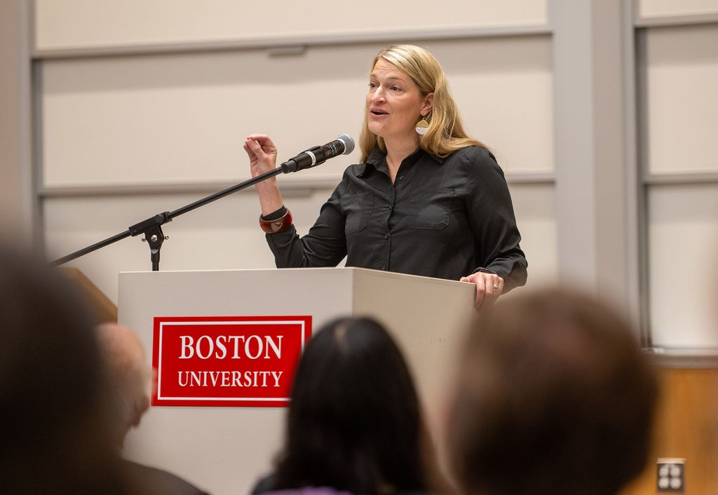 Photo: Maggie Dietz, a white woman with blonde hair and wearing a black collared shirt, speaks into a mic at a white podium with a red "Boston University" sign on it.