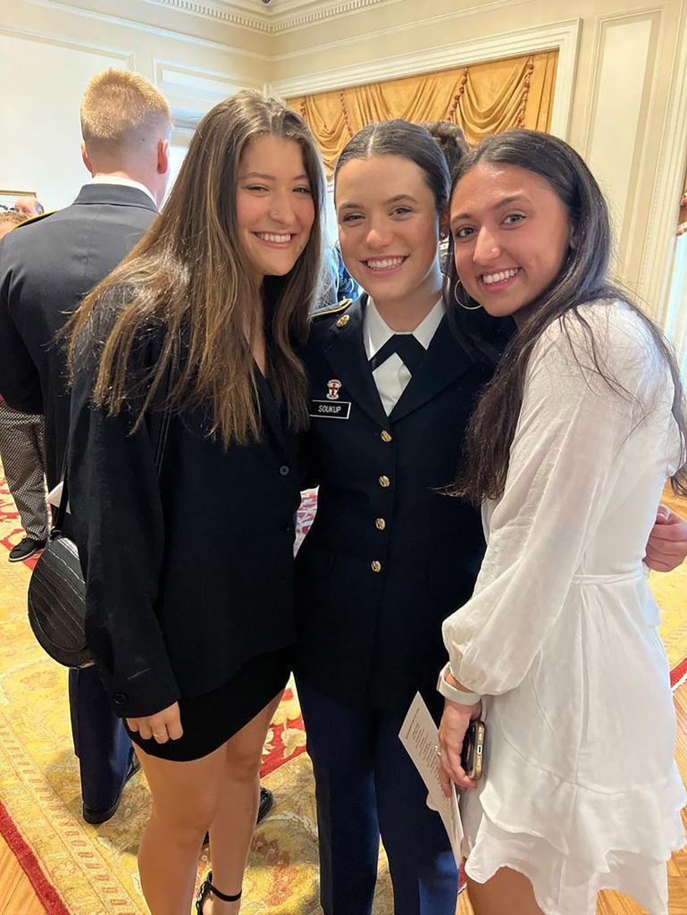Photo: Three young women pose for a photo together. The woman in the center wears a black military style suit, the women on the left wears a black dress, and the woman on the right wears a white dress.