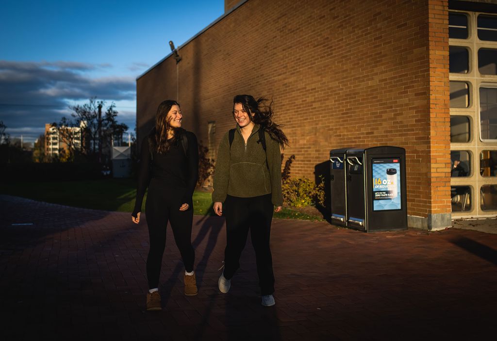 Photo: Two students walking near the West campus dining hall of BU's campus smile and laugh together. They wear casual Fall clothes as the sun rises.
