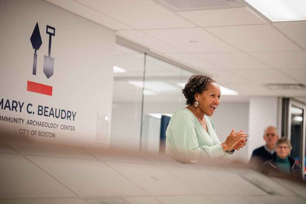 Photo: Rev. Mariama White-Hammond, a Black woman wearing a light green top, smiles as she talks to an unseen audience. She stands in front of a wall that reads "Mary C. Beaudry Community Archaeology Center, City of Boston".