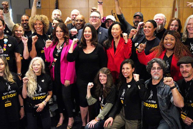 Photo: GFran Drescher, center, joined with National Executive Director and Chief Negotiator Duncan Crabtree-Ireland, and the TV/Theatrical Negotiating Committee members posing with one fist up in the air. They stand in front of a wooden wall.