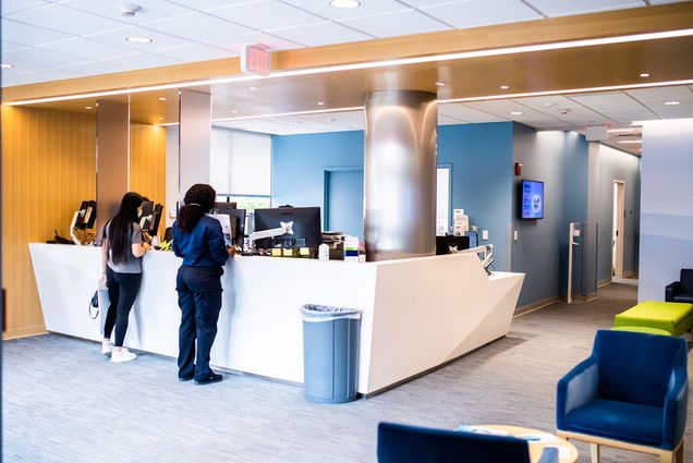 Photo: A stock photo of the updated Student Health Services. Two people stand at a front desk in spacious, clean white and blue waiting area.