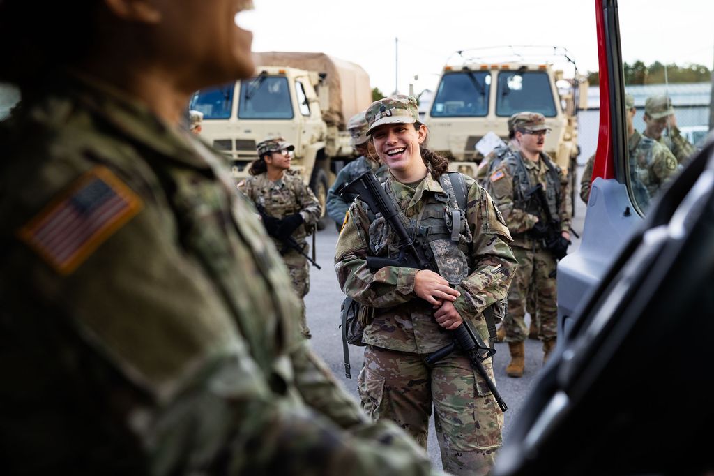 Photo: Audrey Soukup, a young white woman wearing ROTC army fatigue gear and holding a alrge rifle, laughs at something the person in the foreground is saying. The person in the foreground sits in the driver seat of a van and laughs. Other students in similar gear can be seen mingling behind Soukup.